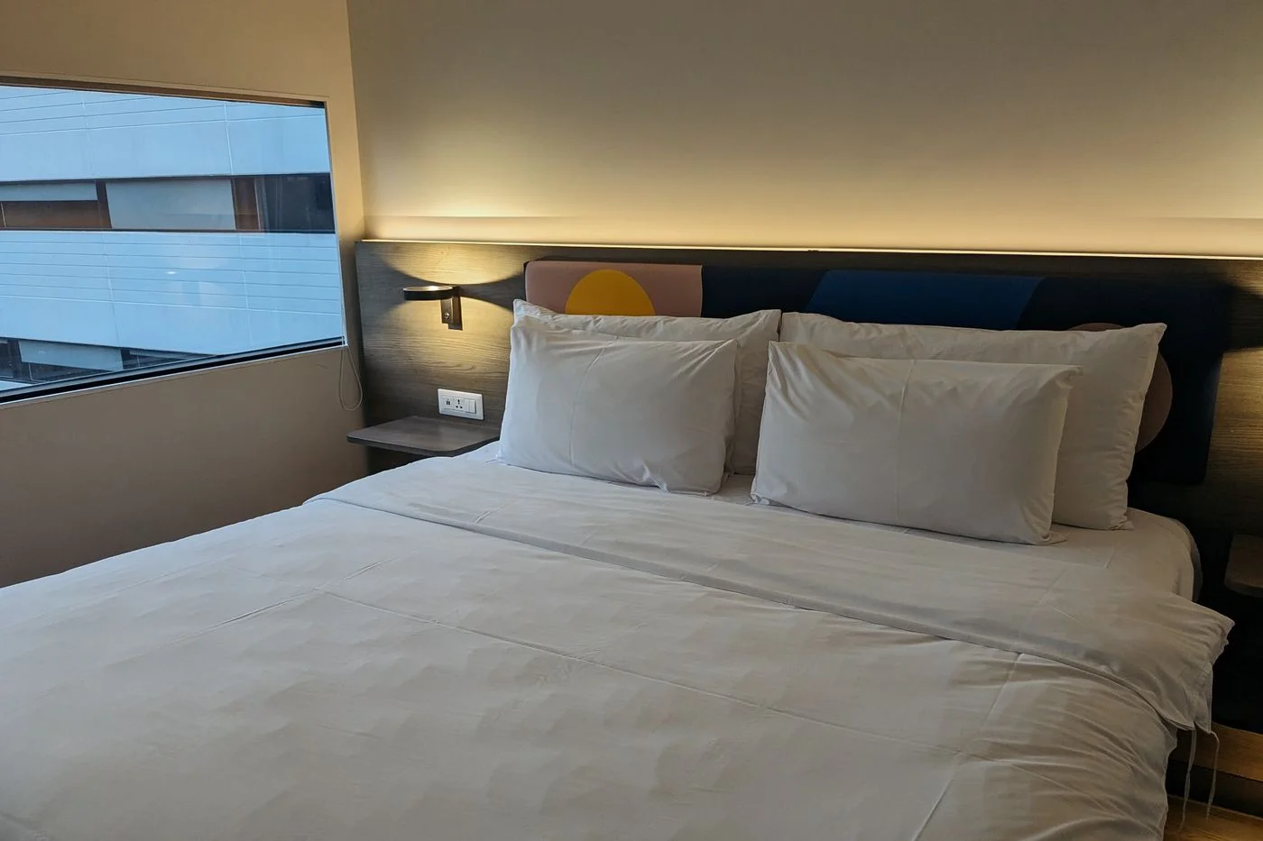 A hotel bed beside a window overlooking nearby buildings